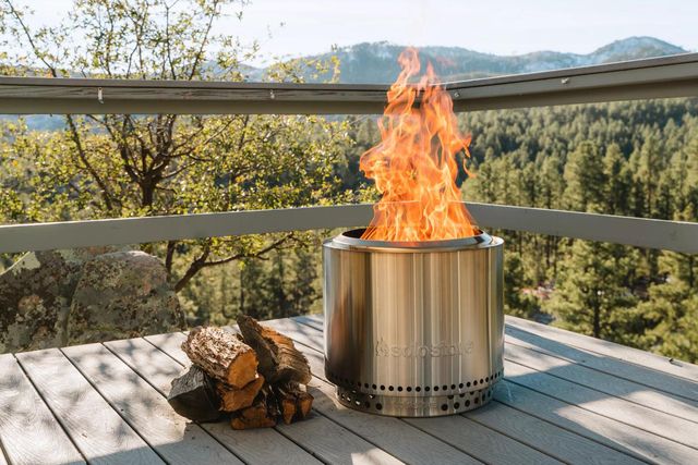 a solo stove with fire on a deck next to chopped wood overlooking a mountain range